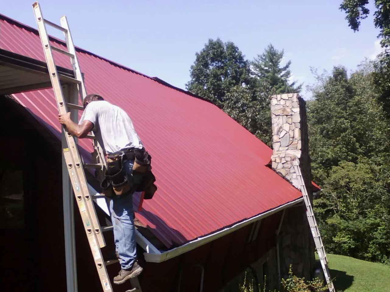 Roofer on a ladder beside a red metal roof and stone chimney — Soffit & Fascia Repair by Roofing Brothers in Alvarado